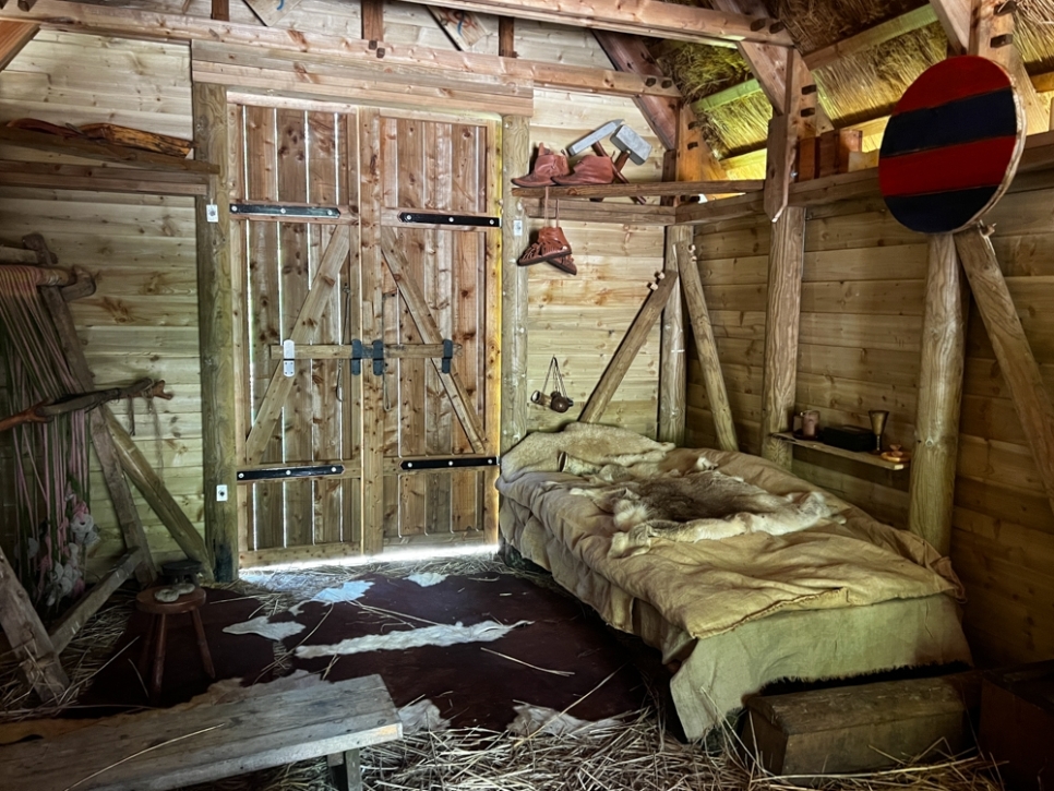 Picture shows inside the longhouse at martin mere. There is a bed to the right, a cow skin rug on the floor, and various pieces of furniture around.